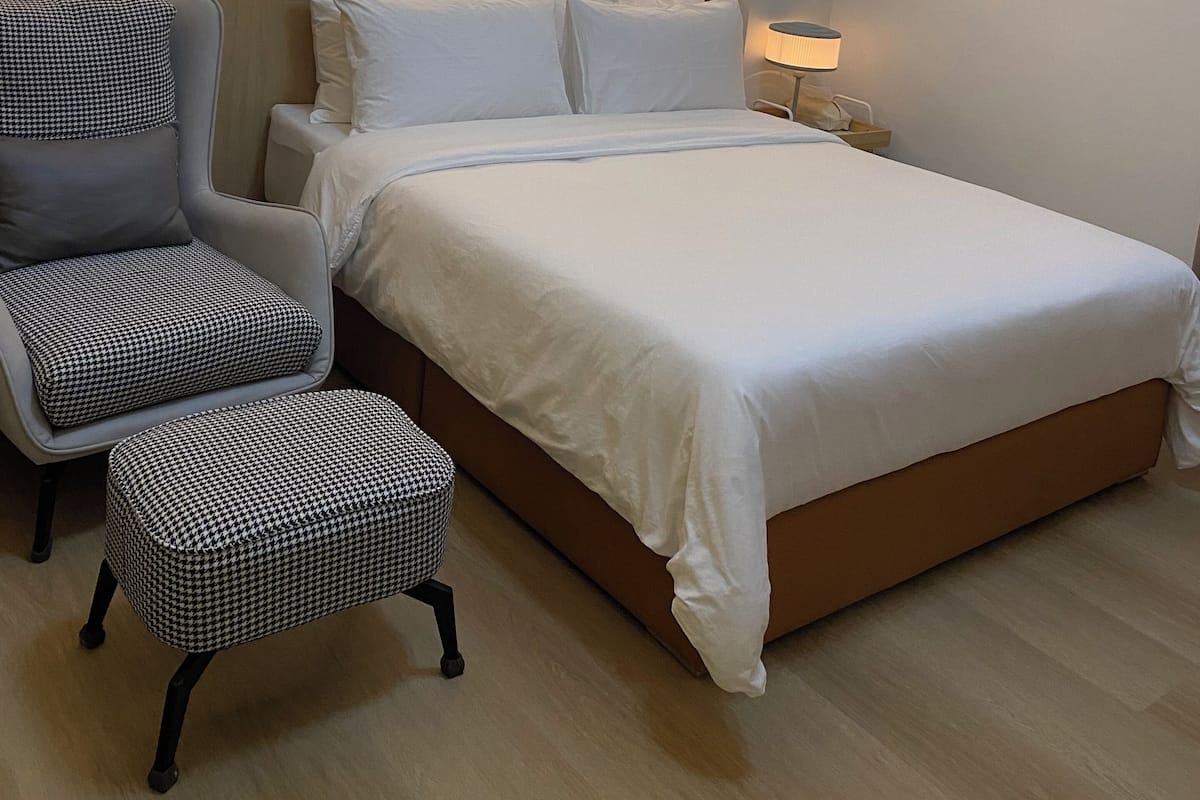 A hotel room with a bed, a chair, an ottoman, a bedside table, and a wall-mounted picture.