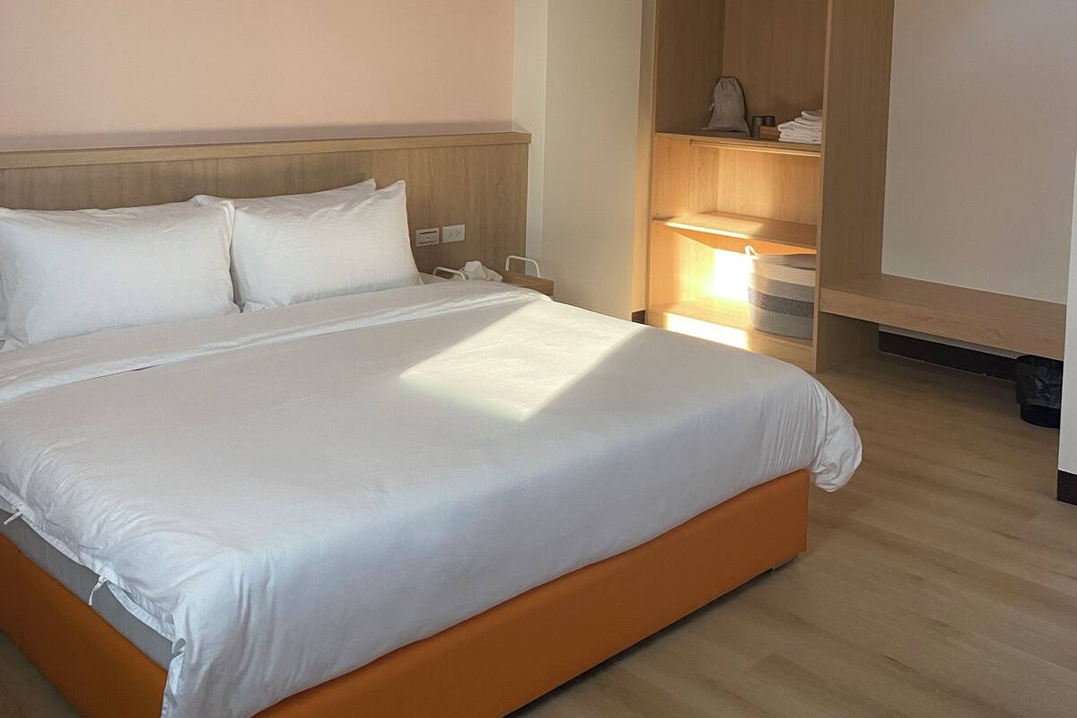 A hotel room with a large bed, wooden headboard, a wardrobe, and a bedside table with a lamp.