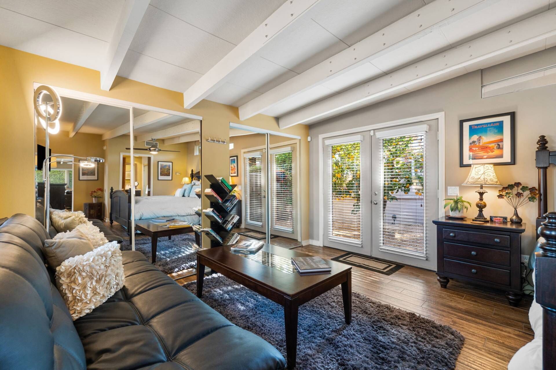A spacious living room with a dark leather sofa, a coffee table, and a bookshelf. There is a sliding glass door leading to a balcony with plants.