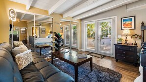 A spacious living room with a dark leather sofa, a coffee table, and a bookshelf. There is a sliding glass door leading to a balcony with plants.