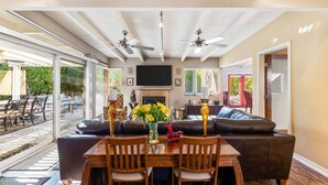 A spacious living area with a large sofa, a dining table with chairs, and a fireplace. There are ceiling fans, a TV mounted on the wall, and a sliding glass door leading to an outdoor patio.