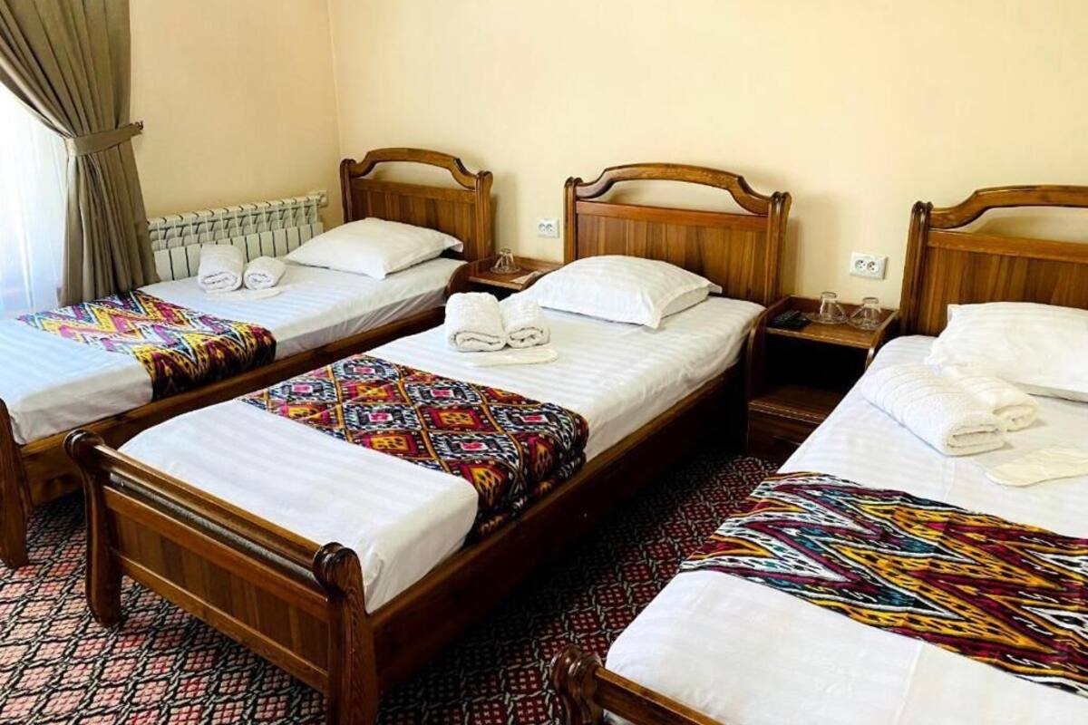 A hotel room with three wooden beds, each with a white mattress and a colorful blanket. There are two bedside tables with lamps, and a radiator under the window.
