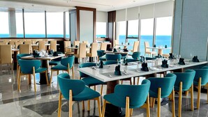 A modern dining area with teal chairs and wooden tables, large windows with a view of the ocean, and a clean, contemporary design.
