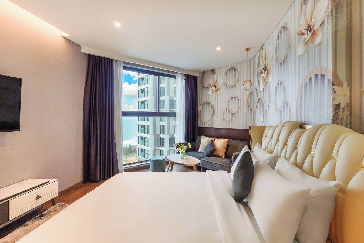 Une chambre d’hôtel avec un grand lit, un canapé, une petite table ornée d’un vase de fleurs et une vue sur les immeubles par la fenêtre.