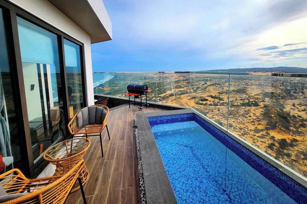 Un balcon avec une piscine, des fauteuils en osier et un barbecue, le tout offrant une vue sur un paysage désertique.