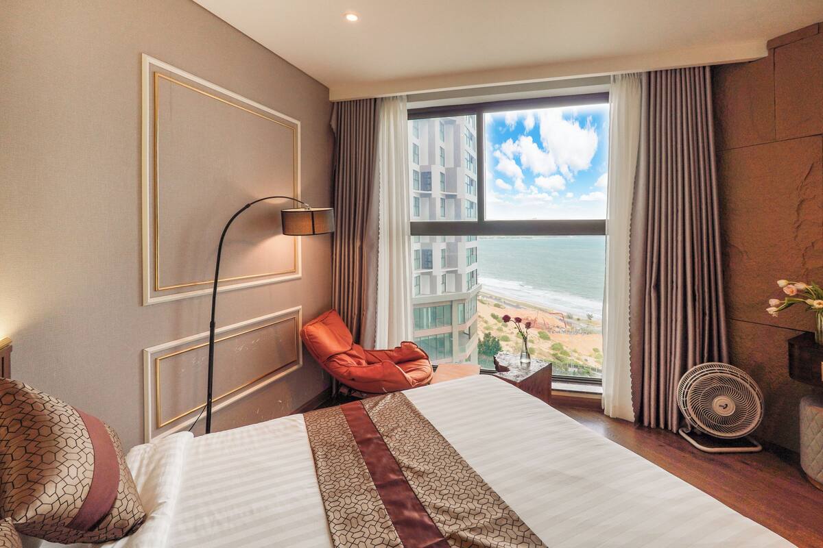 Une chambre d’hôtel avec un grand lit, une chaise, un ventilateur sur pied et une vue sur l’océan par la fenêtre.