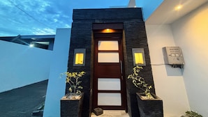 Entrada de un edificio moderno con fachada de piedra oscura, puerta de madera y dos apliques de pared iluminados.