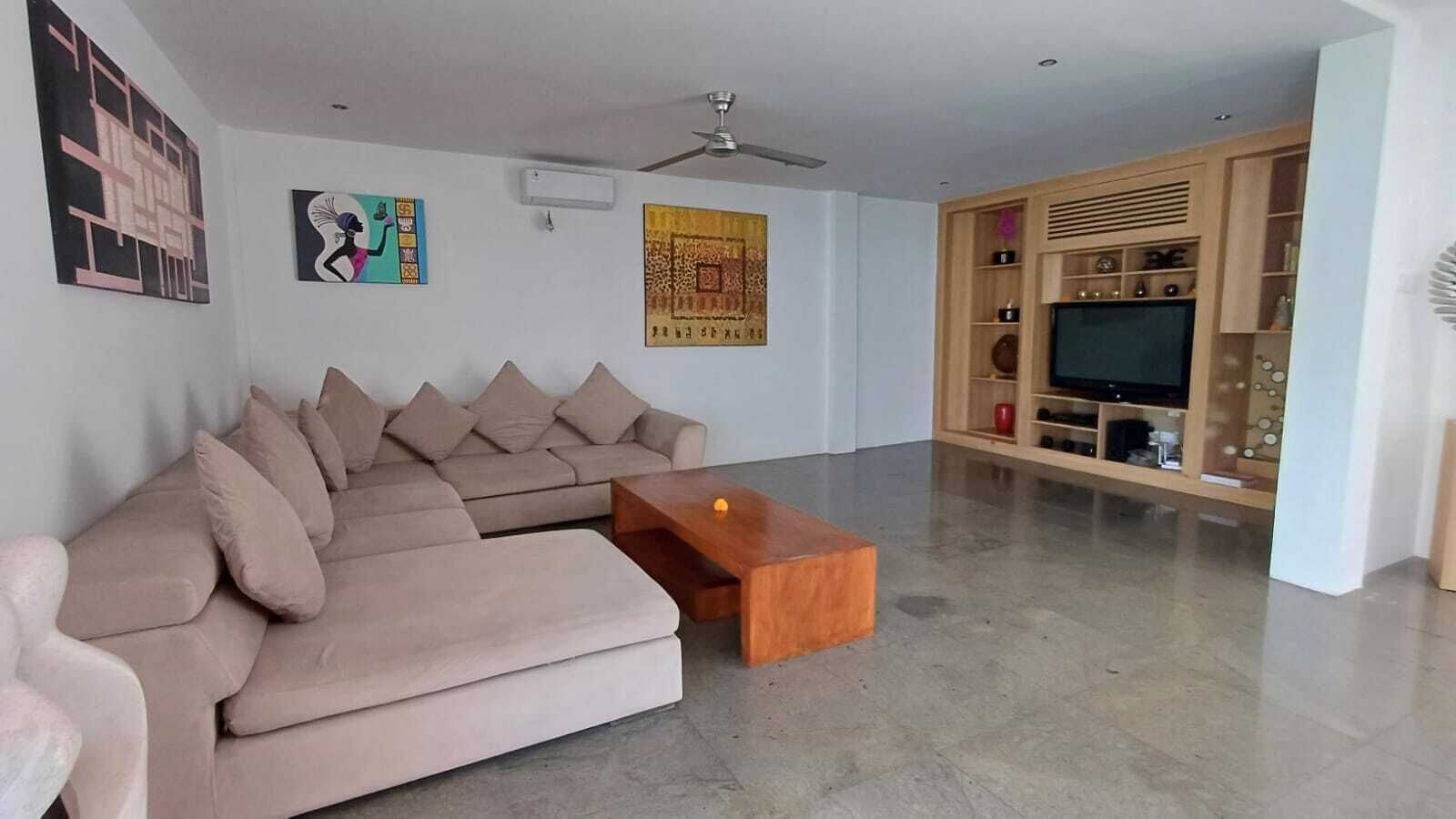 A living room with a beige sectional sofa, a wooden coffee table, a ceiling fan, and a TV stand with shelves.