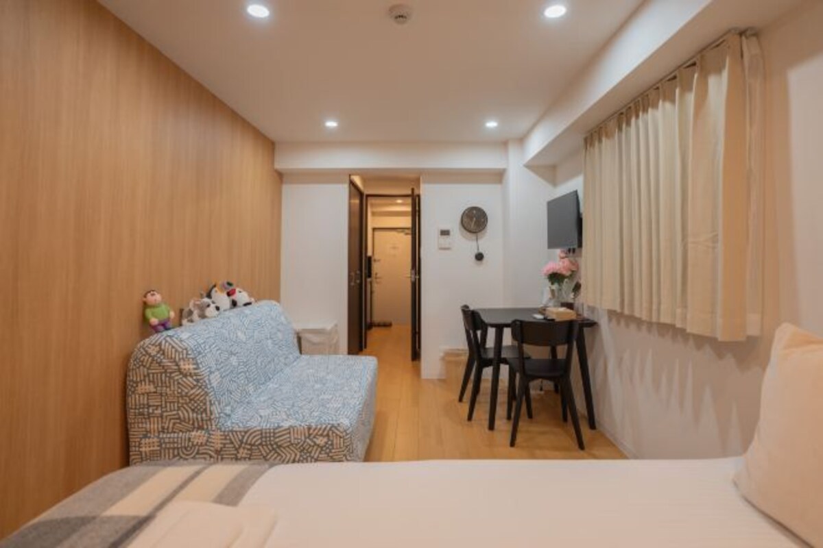 Ein kleines Zimmer mit einem Bett, einem Tisch und einem Stuhl. Eine Holzvertäfelung und ein Fenster mit Vorhängen sind vorhanden.