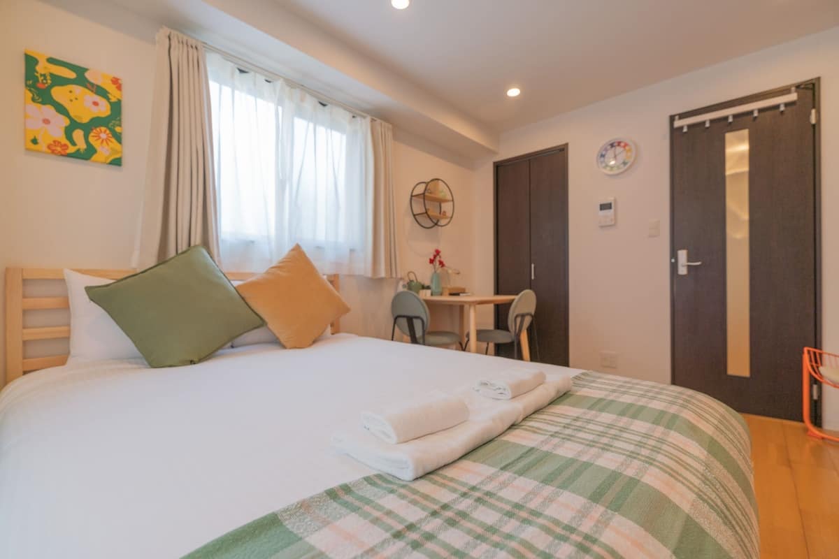 Un lit bien fait, recouvert d’une couverture à carreaux, avec une tête de lit en bois et des oreillers. On y trouve également un bureau avec une chaise et une horloge au mur.