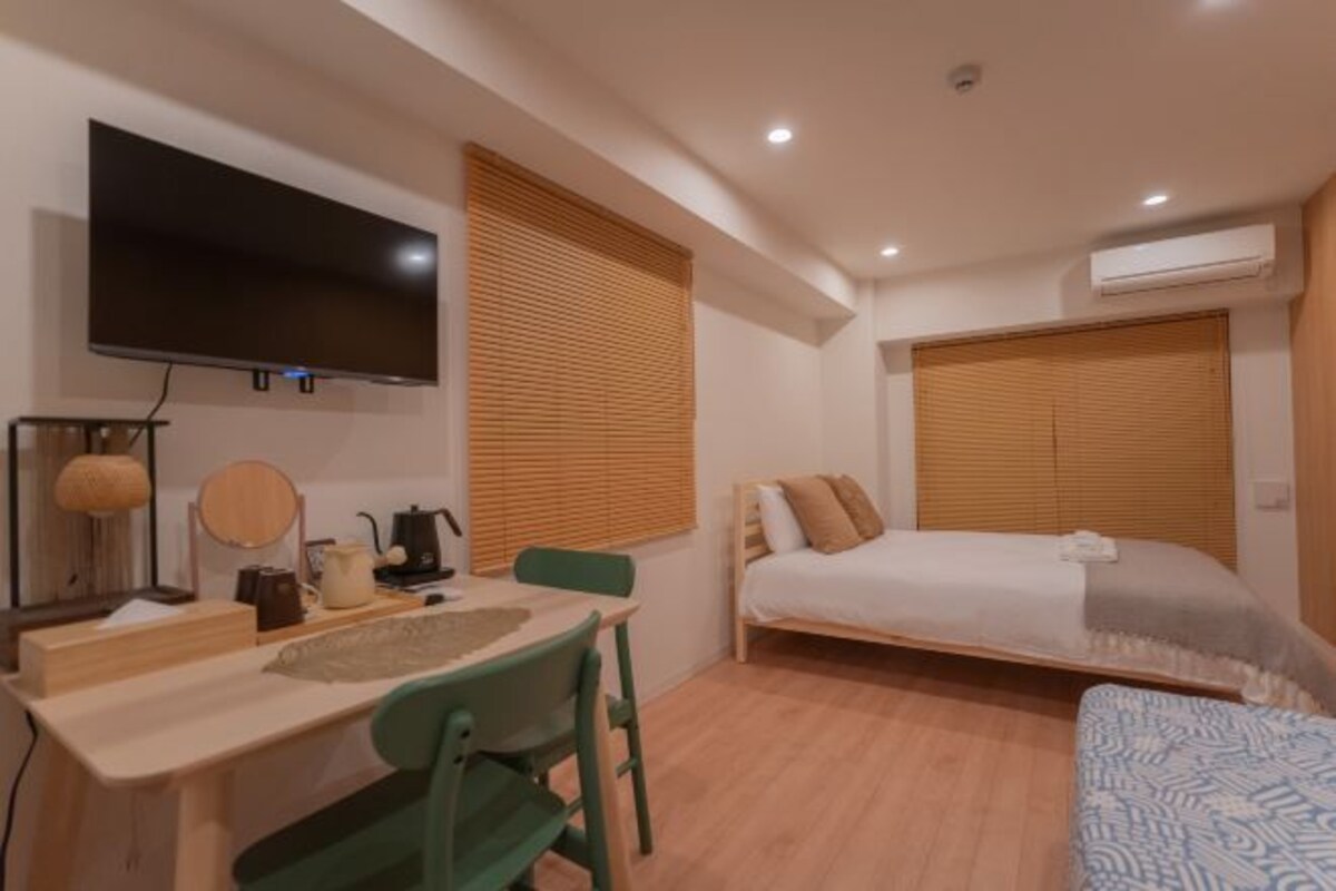 Ein kleines Hotelzimmer mit Bett, Schreibtisch, Stuhl, Fernseher und einem Fenster mit Jalousien.