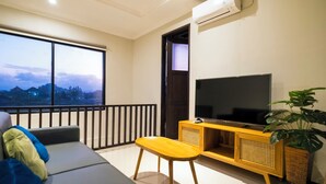 A modern living room with a sofa, a small table, a TV on a wooden cabinet, and a large window offering a view of the cityscape. - Casa Purnama Villa (Sukawati)