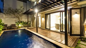 A modern outdoor pool area with a wooden deck, a covered seating area, and a glass door leading to a room with a visible bathroom. - Casa Purnama Villa (Sukawati)