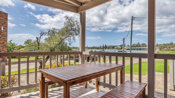 A wooden deck with a table and benches, overlooking a road and water body with trees and buildings in the background.