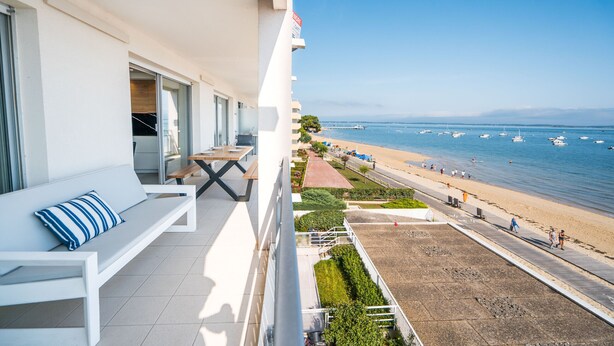 Un balcon avec un canapé blanc et une table, donnant sur une plage, un ciel dégagé et des bateaux à l’horizon.