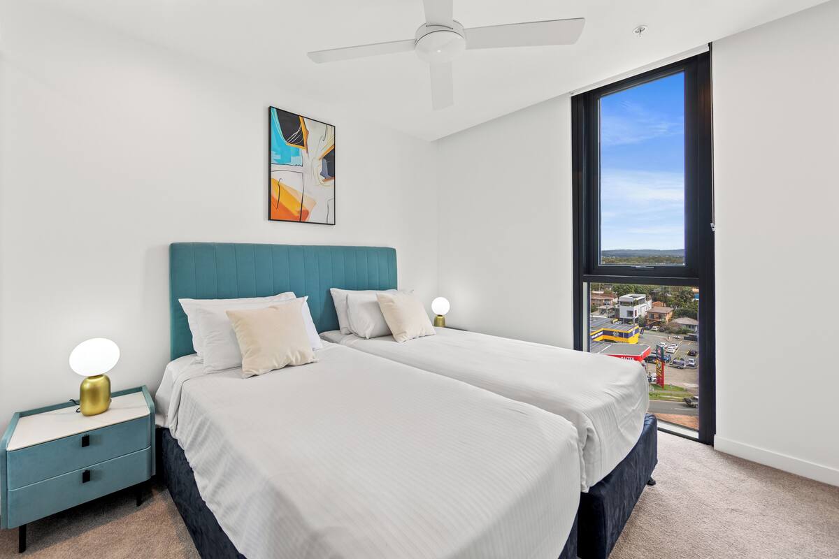 Ein Schlafzimmer mit zwei Betten, einem Deckenventilator, einem großen Fenster mit Blick auf die Stadt und einem Gemälde an der Wand.