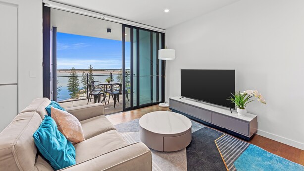 A modern living room with a sofa, a round coffee table, a TV on a cabinet, and a balcony with a view of the water. - Marine Quarter Apartments by CLLIX (Southport)