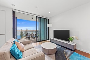 Ein modernes Wohnzimmer mit einer Couch, einem runden Couchtisch, einem Fernseher auf einem Schrank und einem Balkon mit Blick auf das Wasser.