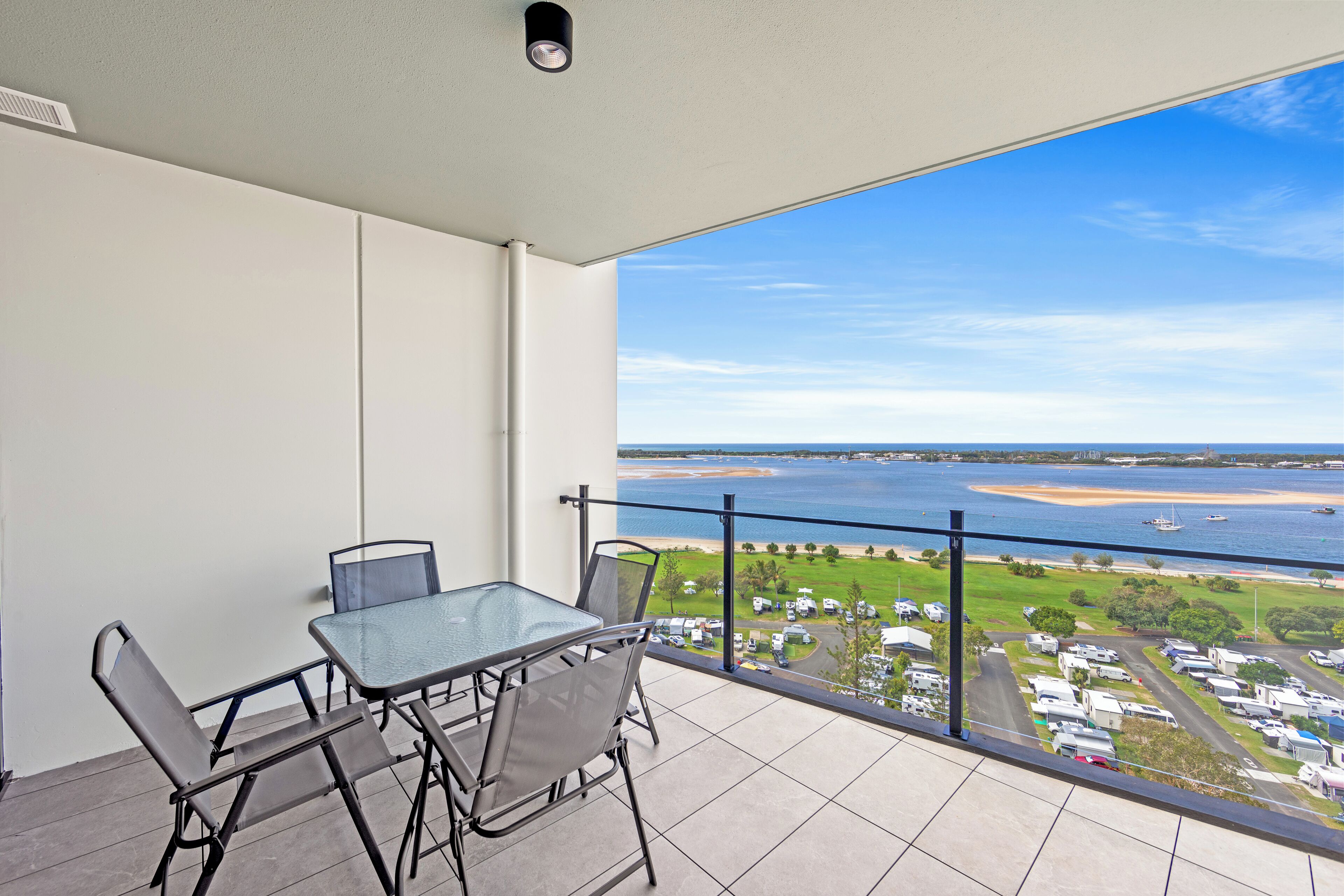 Een balkon met een glazen tafel en stoelen, met uitzicht op een strand en jachthaven.