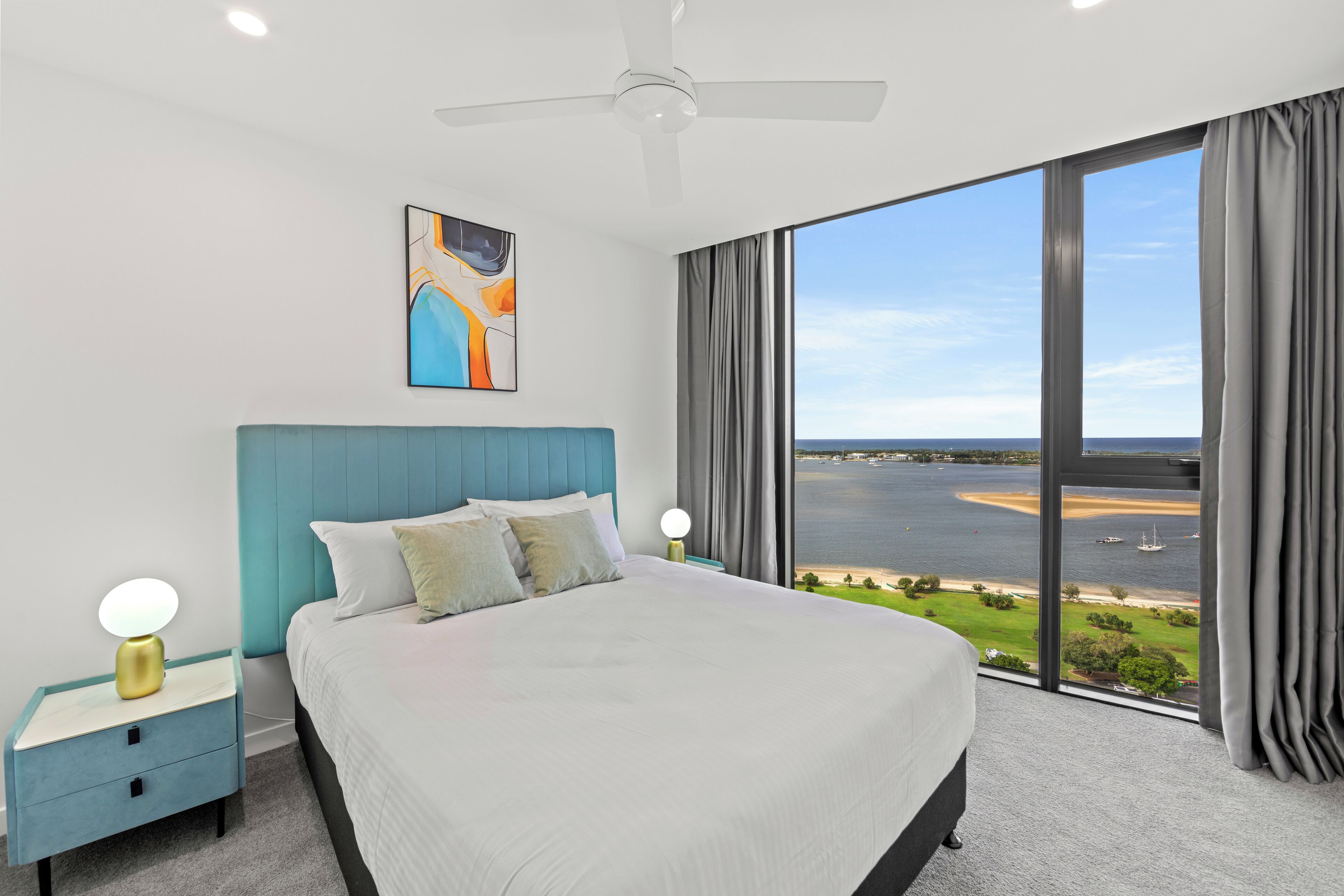 A modern bedroom with a large bed, a ceiling fan, and a view of a body of water through large windows.