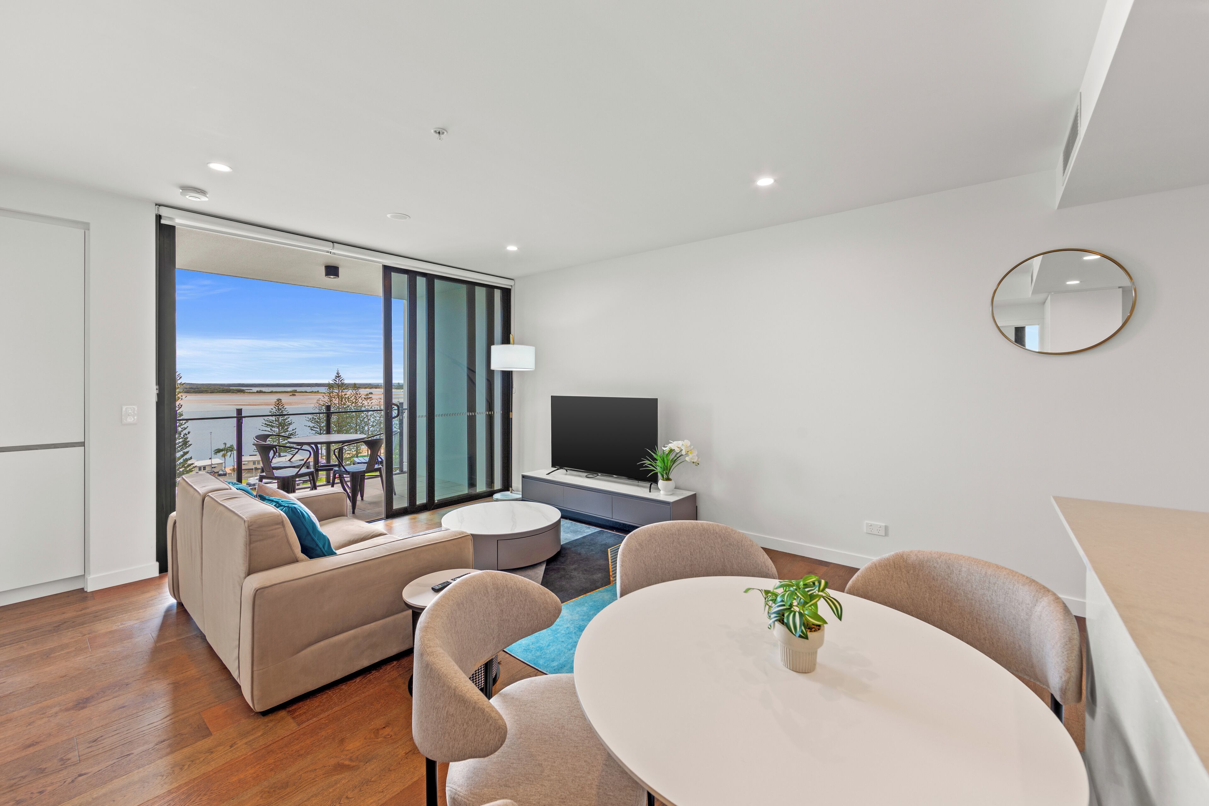 A modern living room with a sofa, chairs, a dining table, a TV, and a balcony with a view.