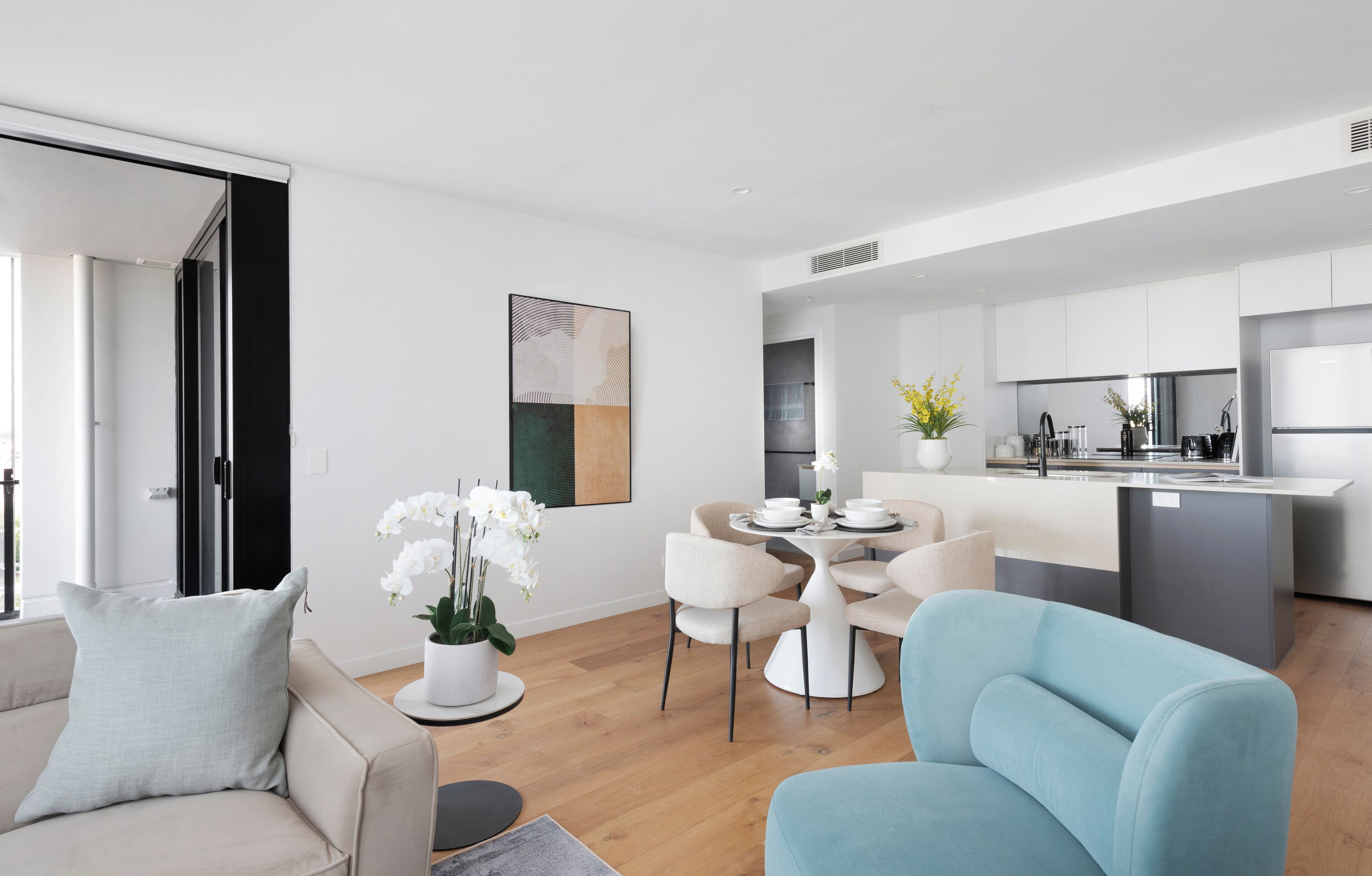 A modern living room with a sofa, a dining table with chairs, and a kitchen area with a refrigerator and cabinets.