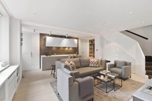 A modern living room with a grey sofa, armchairs, and a coffee table. A kitchen is visible in the background with a white countertop and wooden backsplash. - Knightsbridge & Chelsea Lux 2 bed Duplex (London)