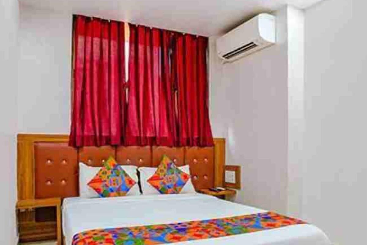 Une chambre d’hôtel avec un lit en bois, des rideaux rouges, une couvre-lit blanche et un petit climatiseur fixé au mur.
