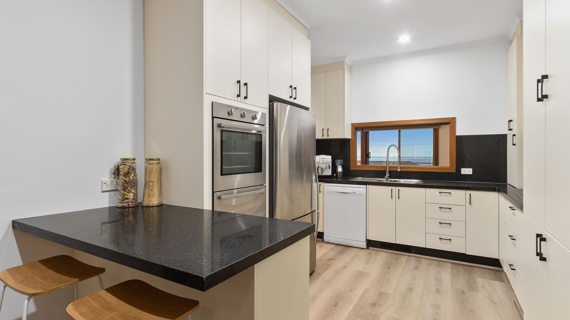 A modern kitchen with stainless steel appliances, a black countertop, wooden stools, and a window with a view.