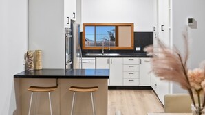 Eine moderne Küche mit Einbauofen, einem Edelstahlkühlschrank und einem Fenster über der Spüle. Auf der Arbeitsplatte stehen zwei Holzbarhocker und eine Vase mit getrockneten Blumen.