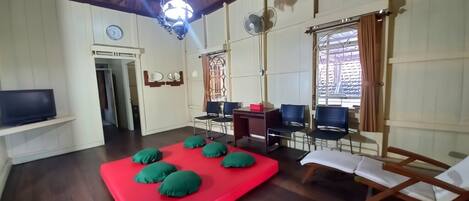 A room with a red mat, wooden flooring, a TV on a shelf, a desk, chairs, and a window with curtains.