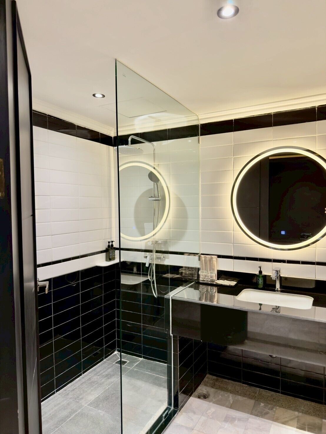 Une salle de bain moderne dotée d’une douche à parois de verre, d’un miroir rond avec éclairage intégré et d’un carrelage noir et blanc.