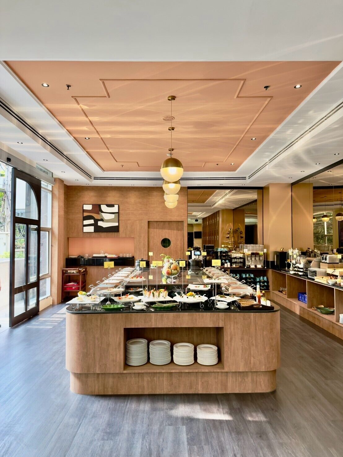A modern hotel lobby with a central buffet area, wooden flooring, and a large window providing natural light.