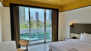 Habitación de hotel con una cama grande, una silla, una mesita de noche y vistas a un paisaje urbano a través de un amplio ventanal.