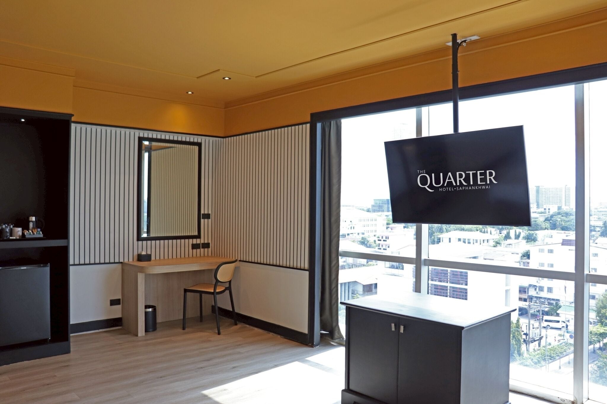 Ein modernes Hotelzimmer mit großem Fenster, einem Schreibtisch, einem Sessel und einem Fernseher, der 'The Quarter Hotel' anzeigt.