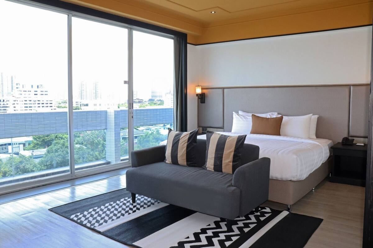 Una habitación de hotel moderna con un ventanal grande, una cama con ropa de cama blanca, un sofá gris y un tapete con estampado blanco y negro.
