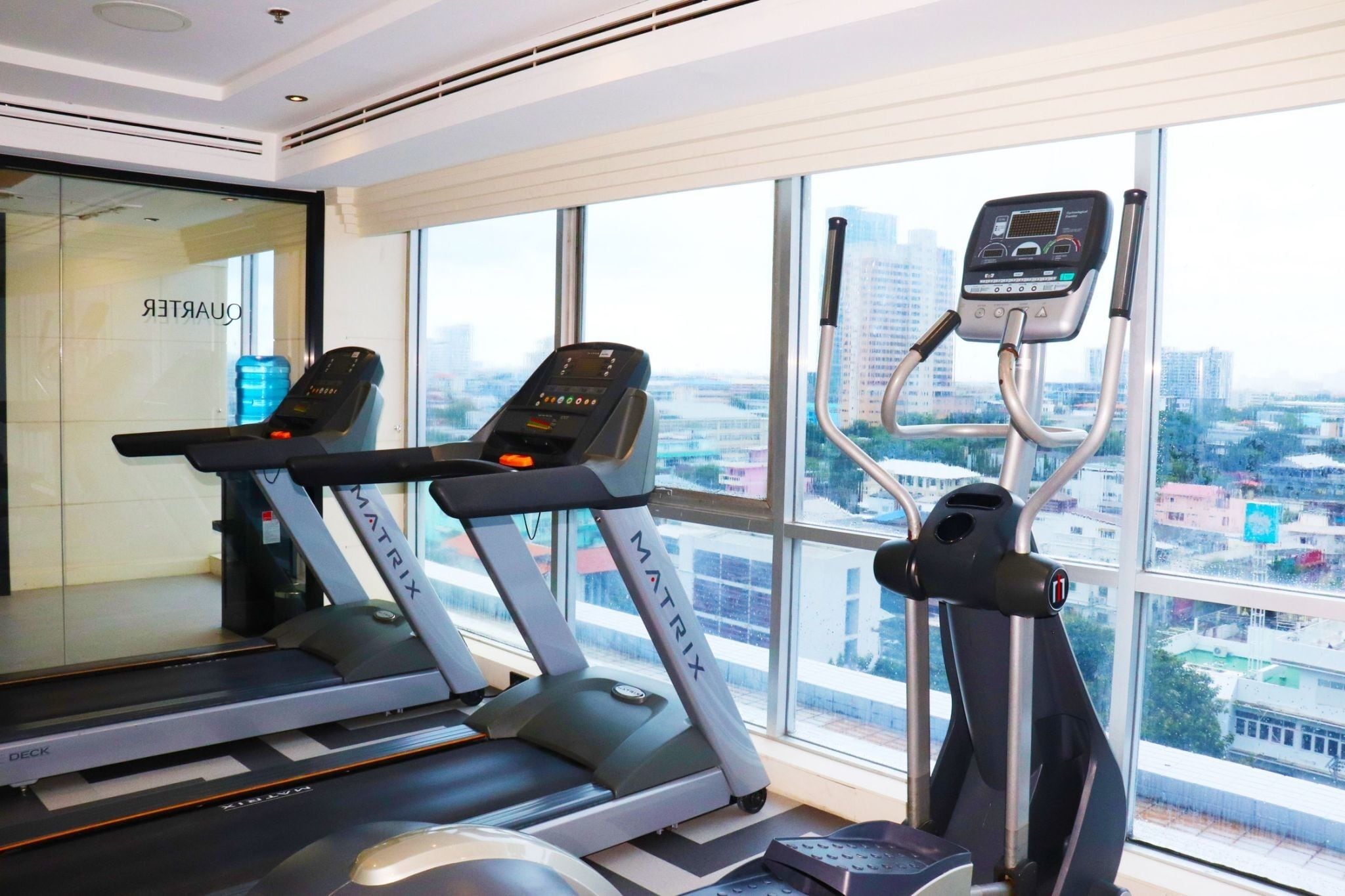 Et fitnesscenter med løbebånd og crosstrainer, store vinduer med udsigt over byen, og en vanddispenser.