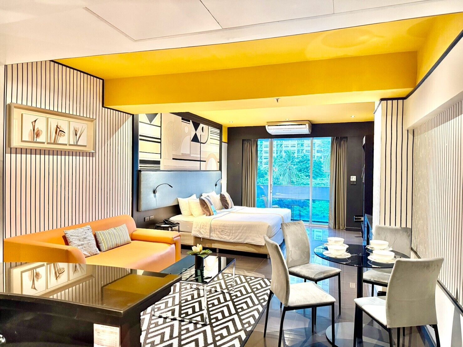A modern hotel room with a bed, a sofa, a dining table, and a view of the outdoors.