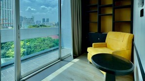 Une chambre moderne dotée d’une grande porte-fenêtre coulissante donnant sur un balcon, d’un fauteuil jaune, d’une petite table noire et d’une bibliothèque intégrée.