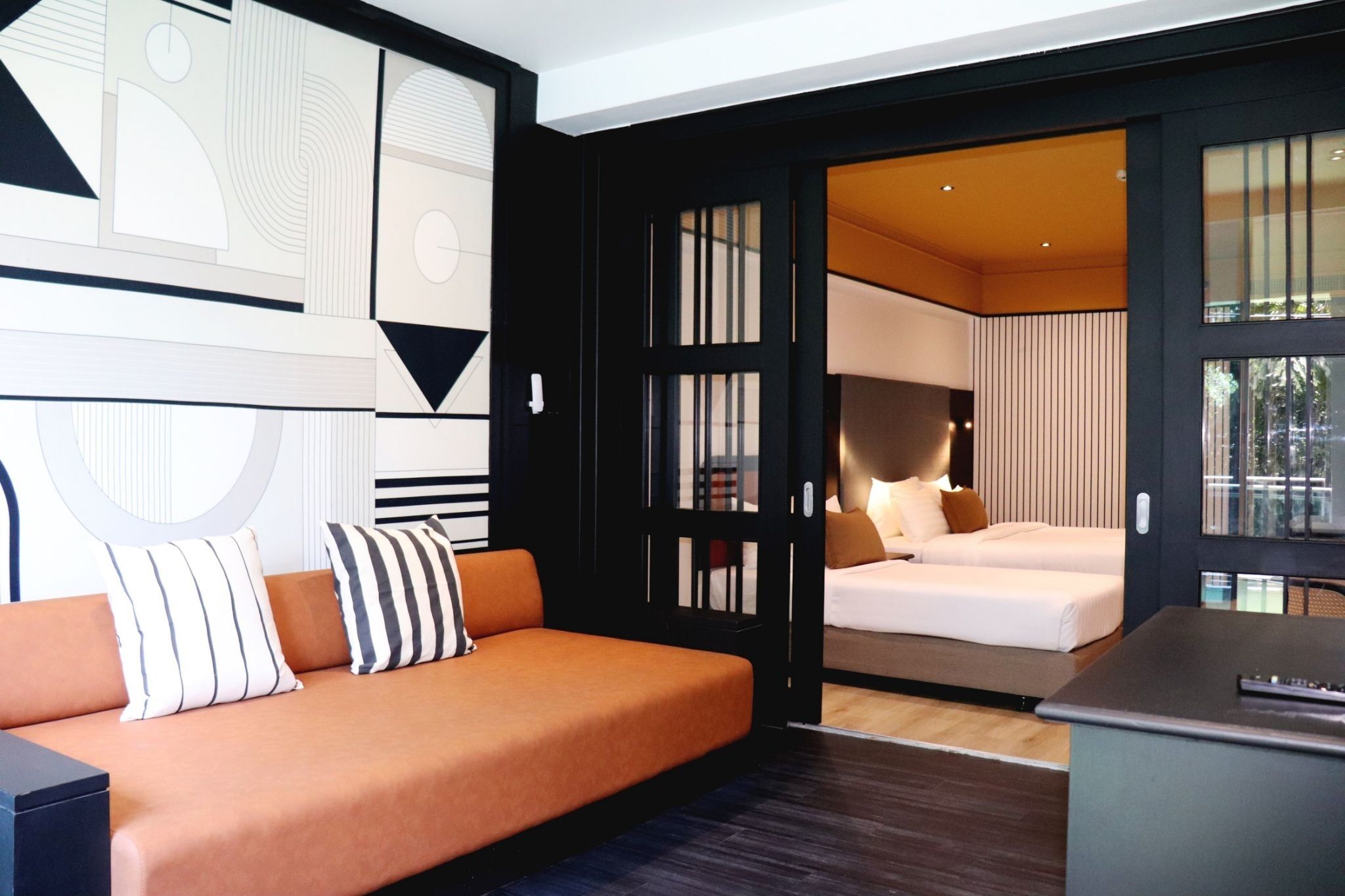 A modern hotel room with a sofa, a bed, and a desk. There is a large window and a door leading to another room.