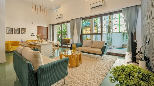 Una sala de estar moderna con un sofá, sillones, una mesa de centro y un televisor. Hay ventanas amplias con cortinas, un ventilador de techo e iluminación colgante.