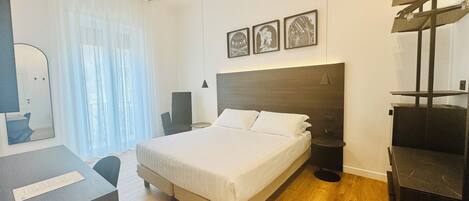 A modern hotel room with a large bed, a desk, a chair, and a mirror. There are three framed pictures on the wall above the bed.