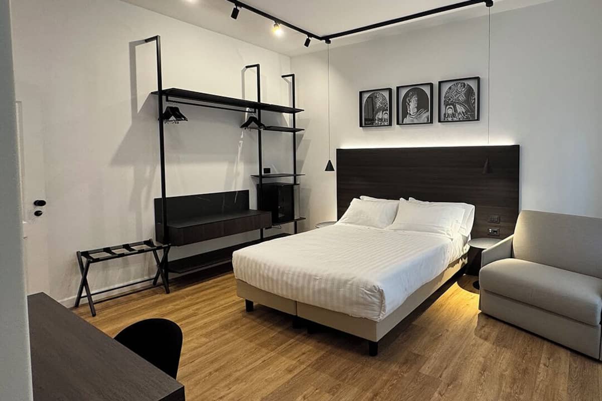 A modern hotel room with a bed, a chair, a desk, and a wardrobe.