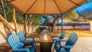 An outdoor patio with a table and chairs under a canopy, surrounded by trees and a house in the background.