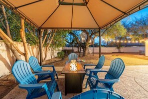 An outdoor patio with a table and chairs under a canopy, surrounded by trees and a house in the background.