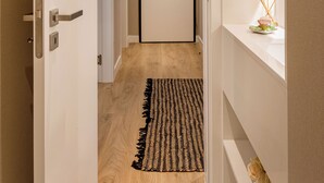 A hallway with a wooden floor, a black and white patterned rug, a white door at the end, and a white wall with a mirror and a small table with a vase and candles. - Cascais Seacoast Premium Apartment (Cascais)
