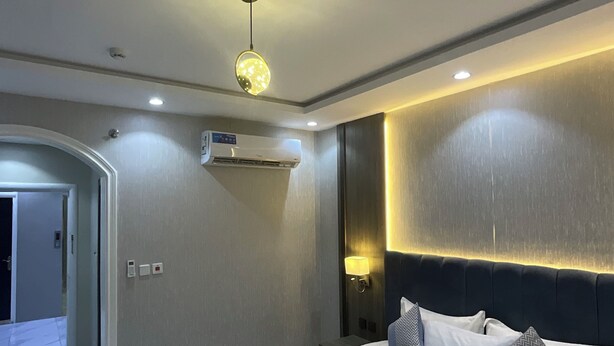 A modern hotel room with a large bed, a wall-mounted air conditioner, a ceiling light fixture, and a door leading to another room.