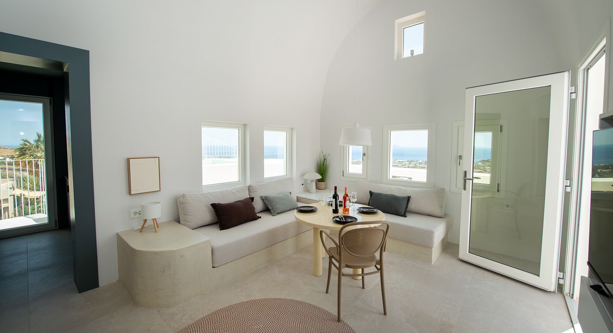 Una sala de estar moderna con un sofá, una mesa de comedor y una silla. Varias ventanas ofrecen vistas al exterior.