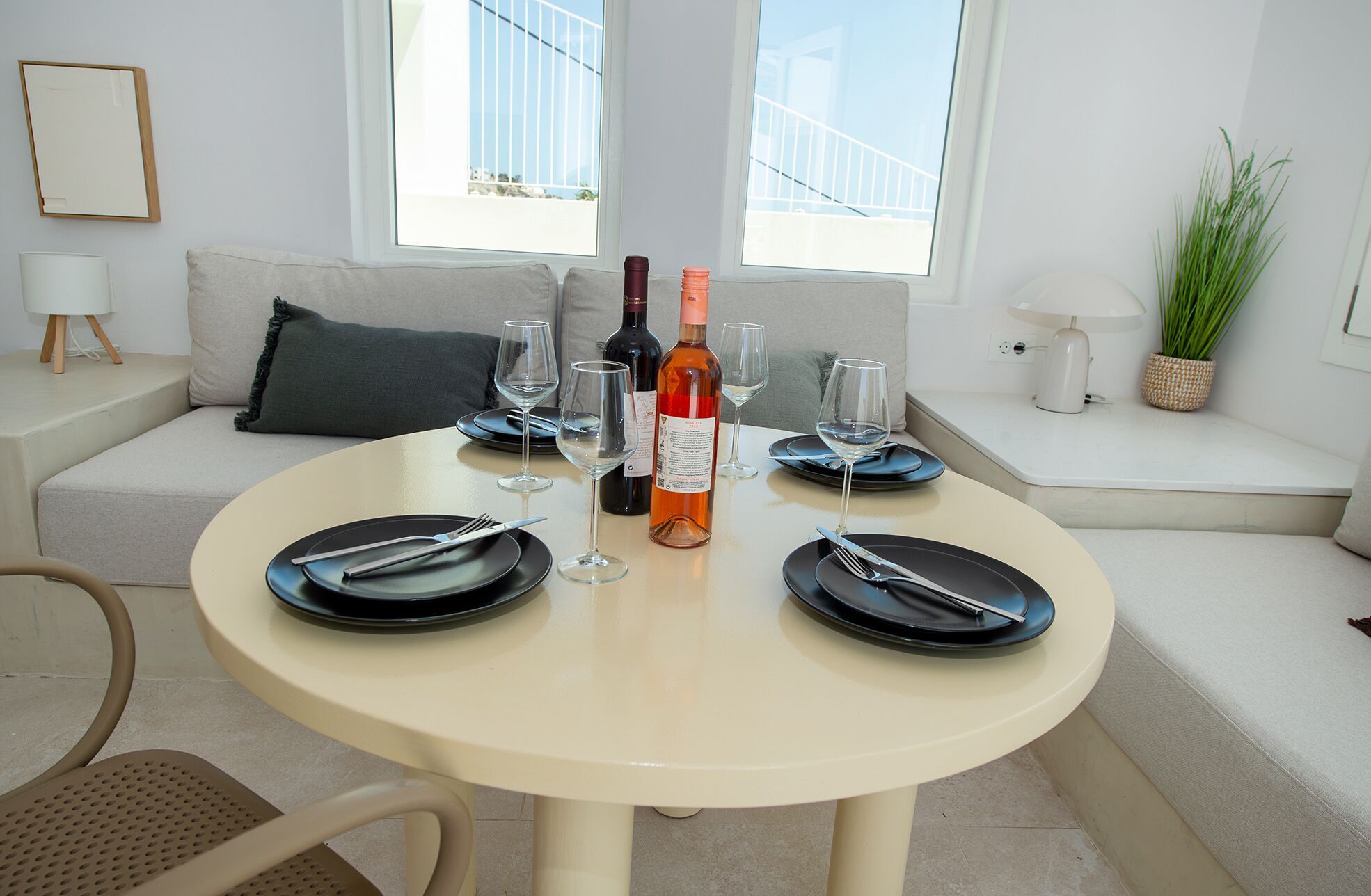 Un comedor con una mesa redonda dispuesta para cuatro personas, con platos negros, copas de vino y cubiertos. Una botella de vino se encuentra en el centro. La habitación presenta un diseño moderno, con un sofá, una planta en maceta y vistas a través de amplias ventanas.