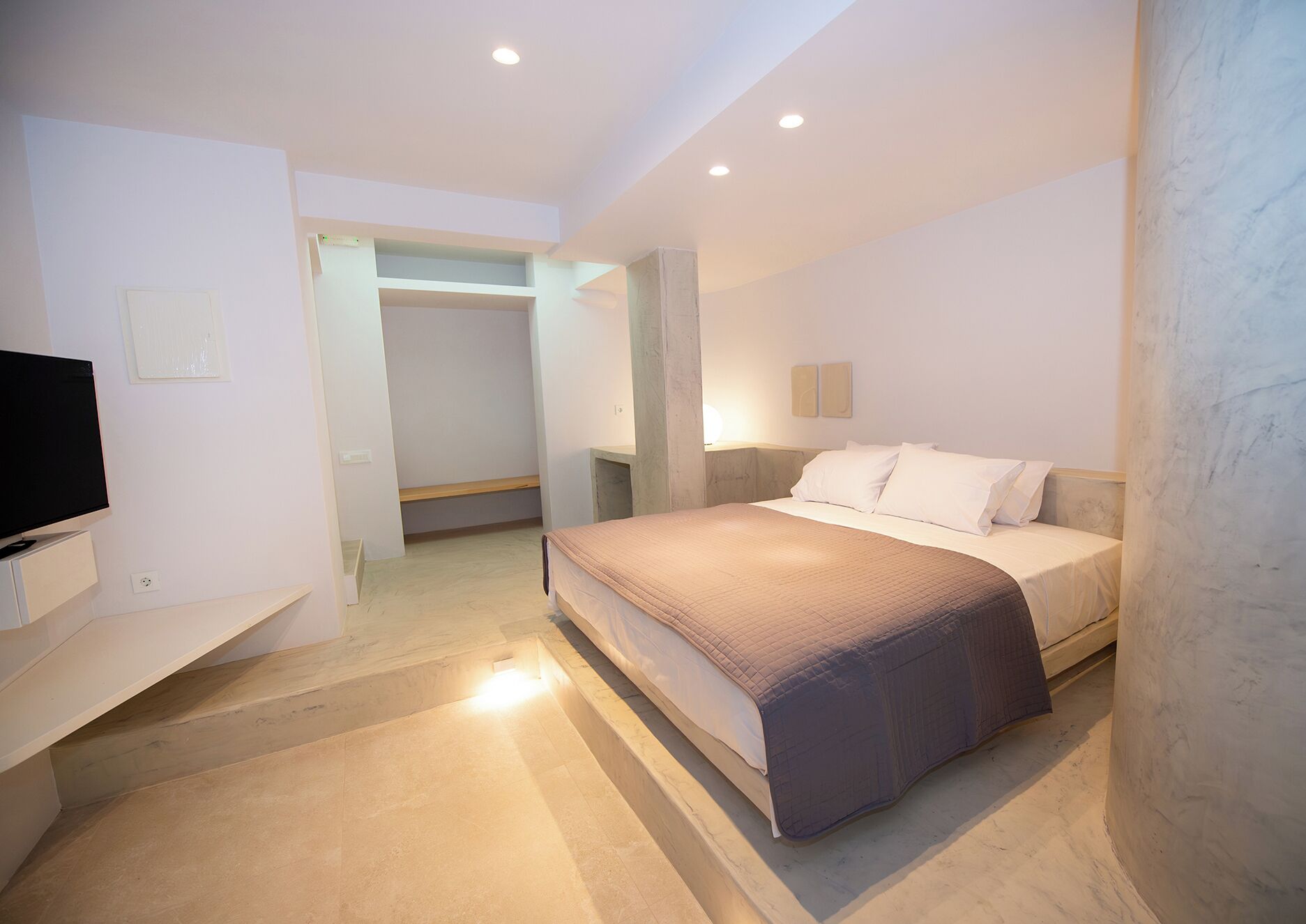 Quarto de hotel moderno com cama, televisão, guarda-roupa e banheiro visível por uma porta aberta.